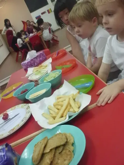 Children seated beside plates and bowls of food on a classroom table.