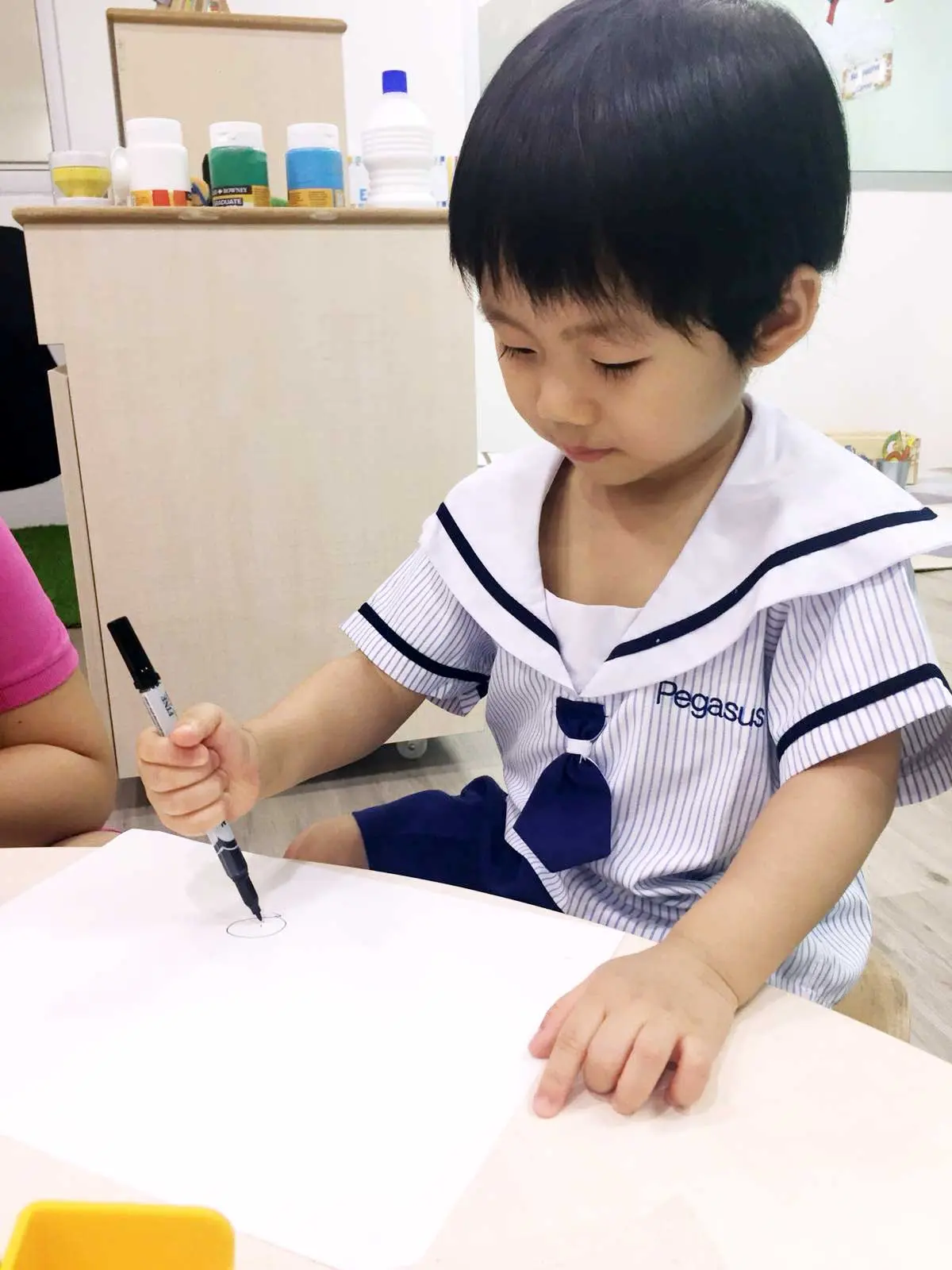 Young child making a circle with a marker at a classroom table.