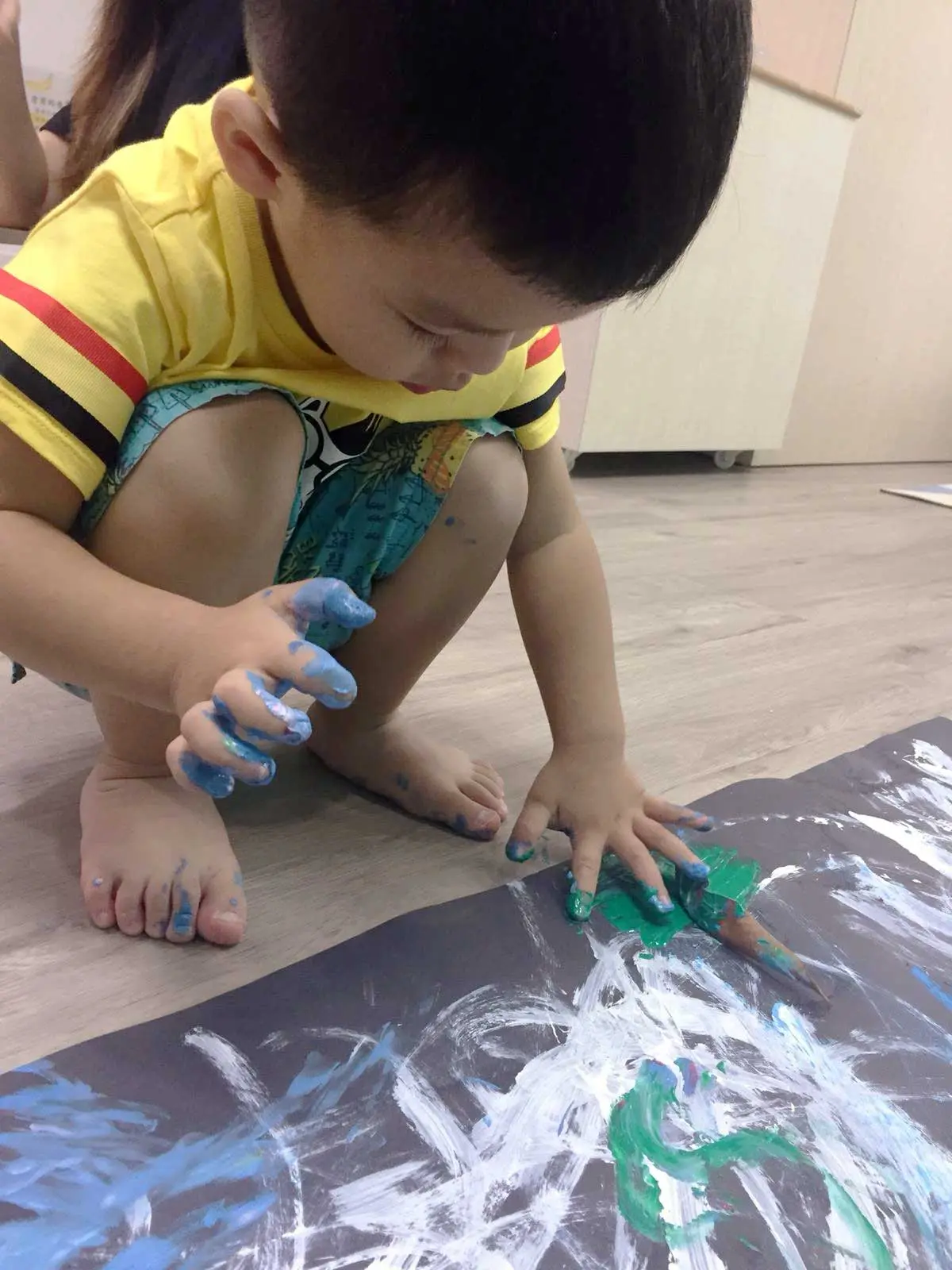 Young child spreading blue and green paint with fingers.