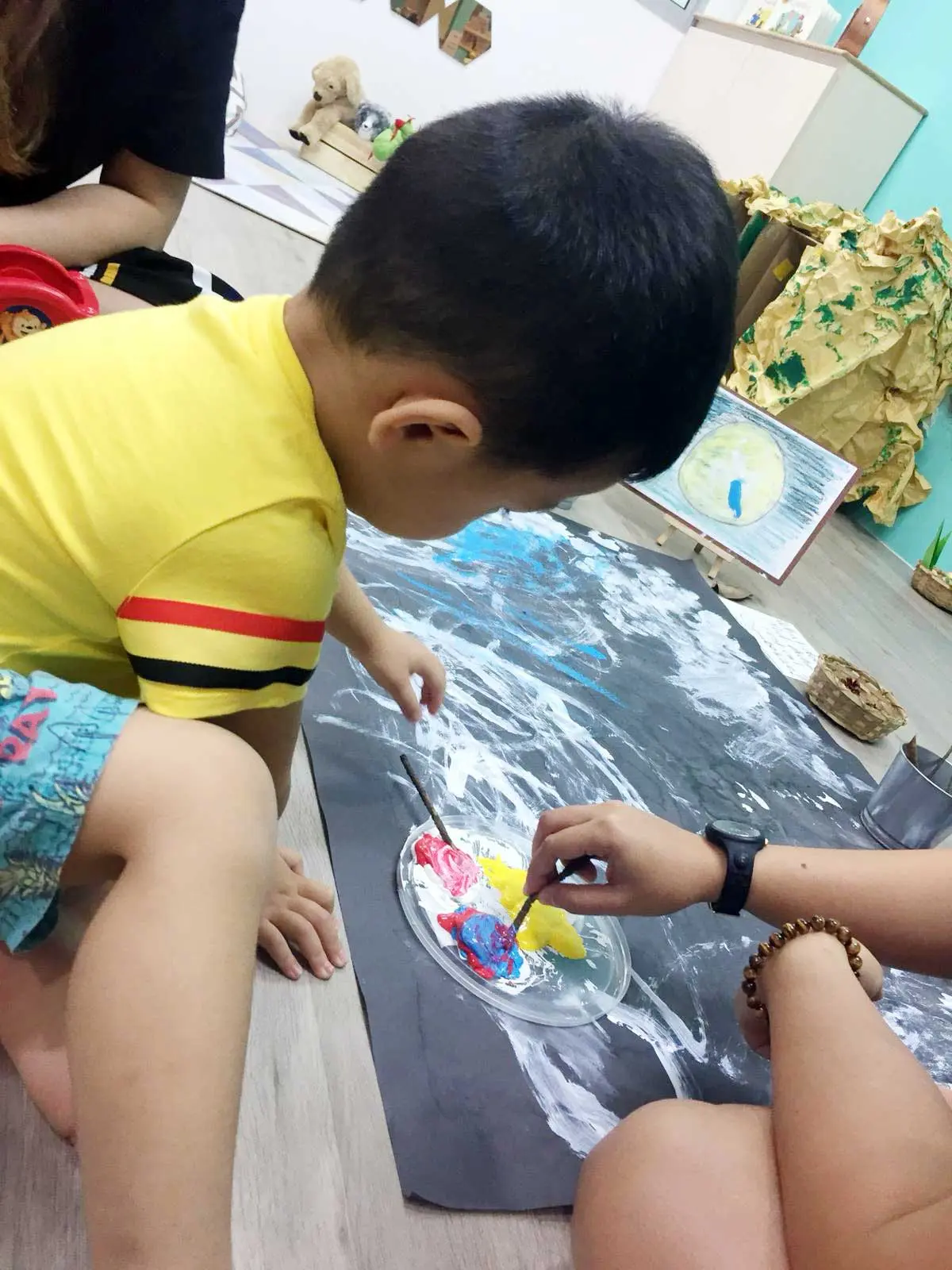 Child watching an adult mix paint beside a large artwork.